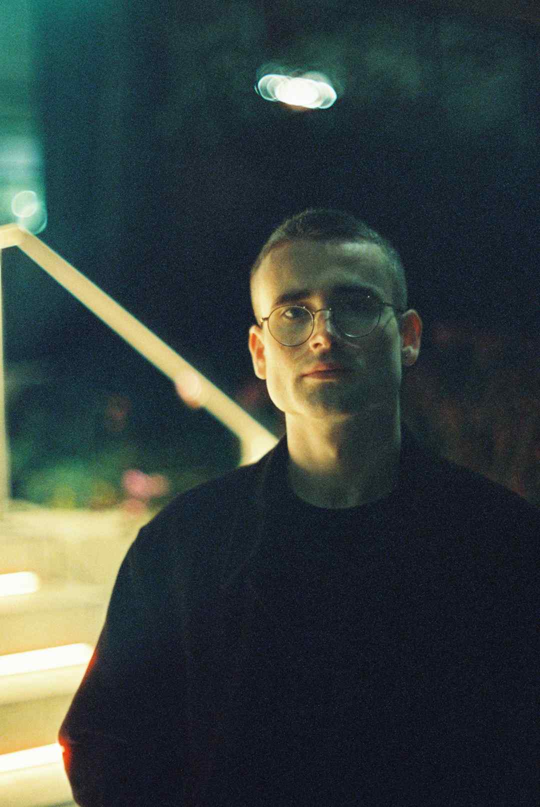 A low-lit film photo of Samuel Powell standing outside the entrance to a building at night.
